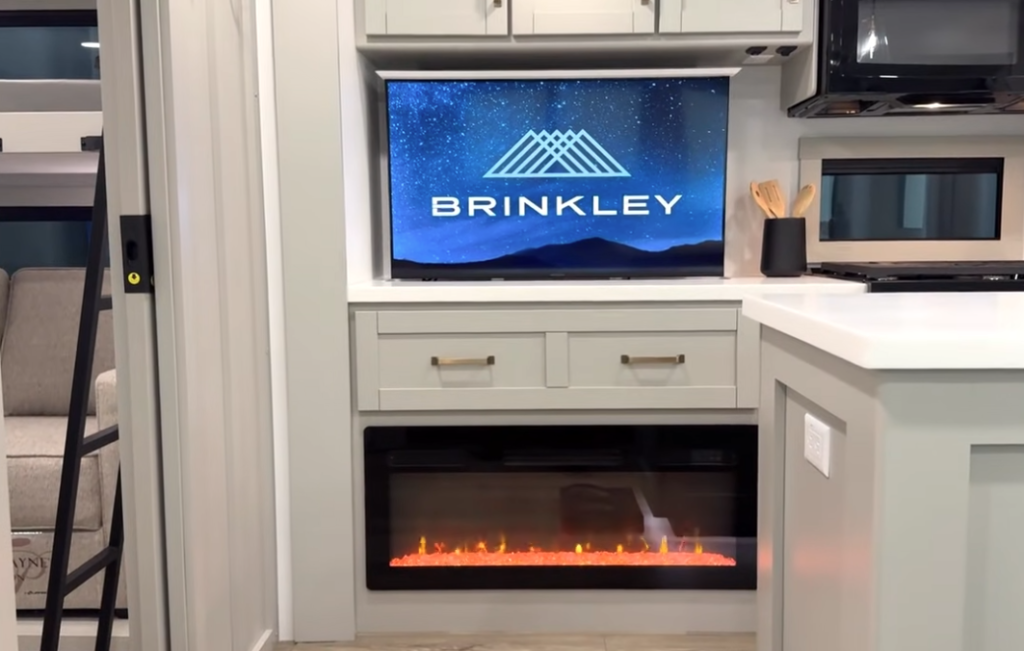 RV entertainment center with a Brinkley HDTV above gray shaker cabinets and an electric fireplace, featuring modern kitchen accents and a bright countertop island.