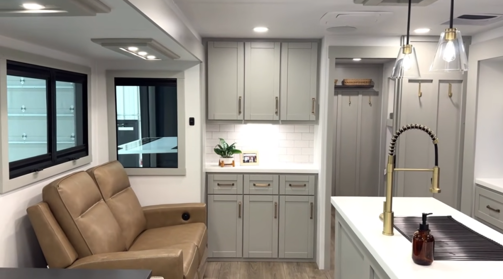 Modern RV interior kitchen with gray shaker cabinets, subway tile backsplash, brass gooseneck faucet, center island, pendant lighting, and tan theater seating in a bright open-concept layout.
