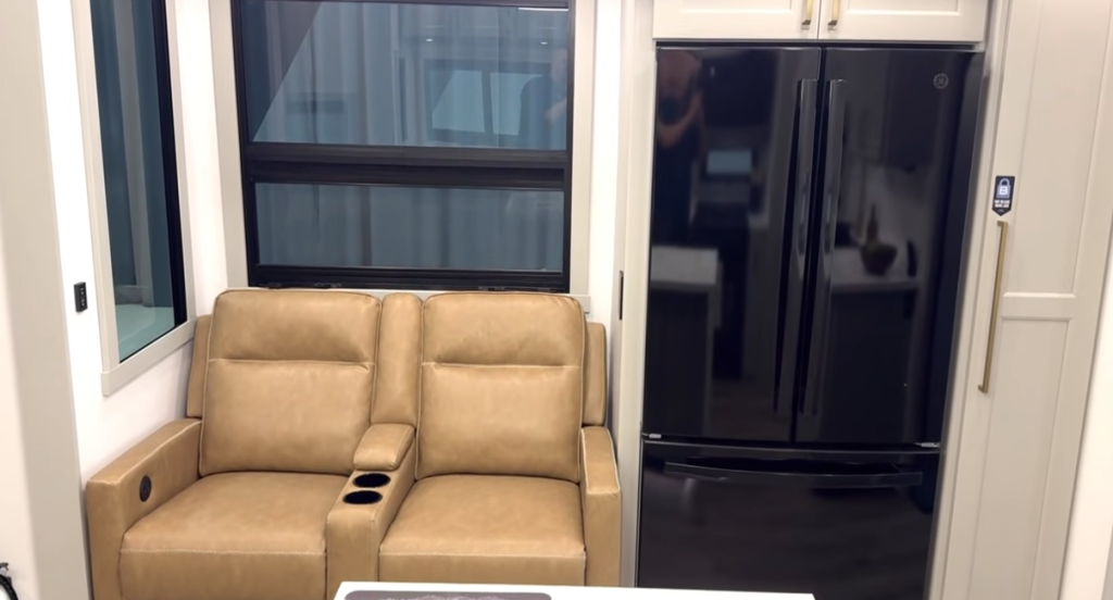 Modern RV interior featuring a tan dual reclining loveseat with cupholders positioned beside a large black French-door residential refrigerator.