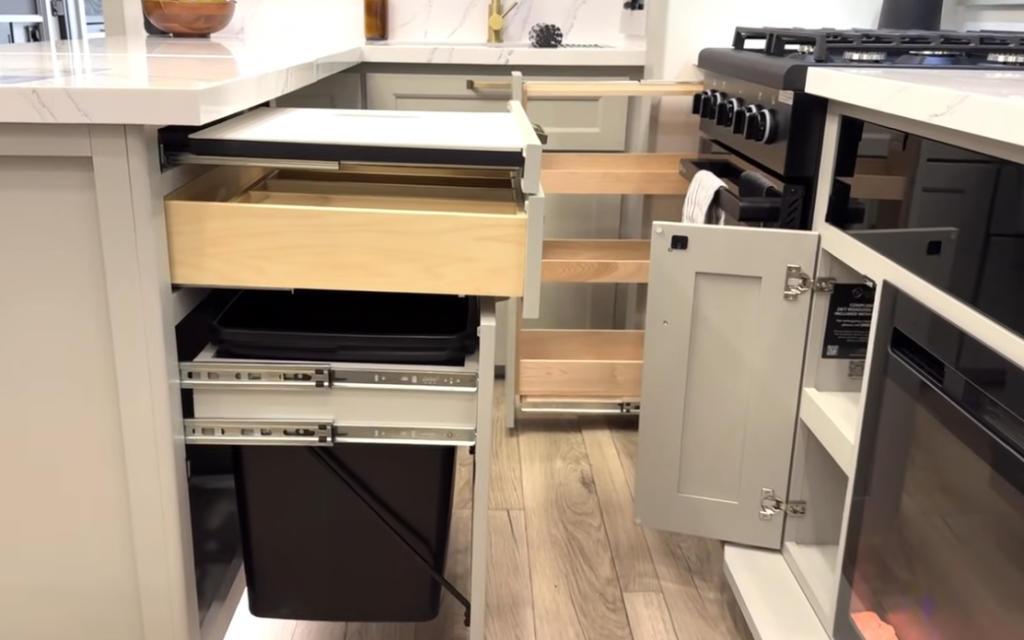 RV kitchen storage system with pull-out trash bins, sliding countertop extension, deep wooden drawers, and pantry racks open beside a modern gas stove in a luxury fifth wheel.