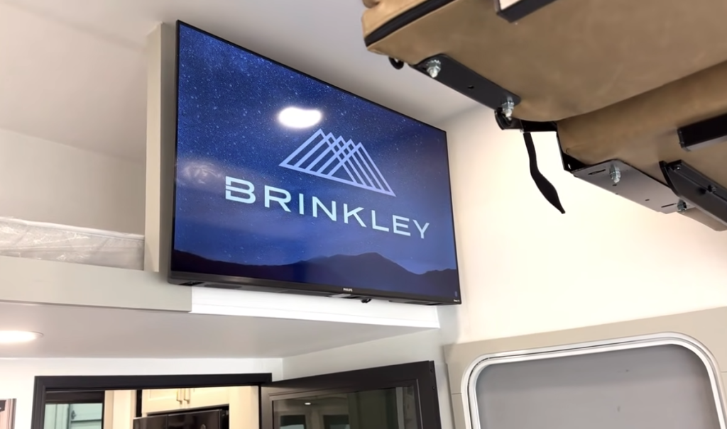 Mounted TV in a Brinkley RV garage displaying the Brinkley logo, shown next to the loft bed and entry door for added entertainment space.