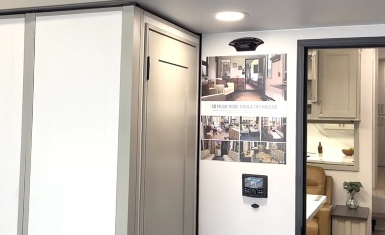 Interior view of a Brinkley toy hauler showing the corner storage cabinet, wall-mounted photo display, and doorway leading into the main living and kitchen area.