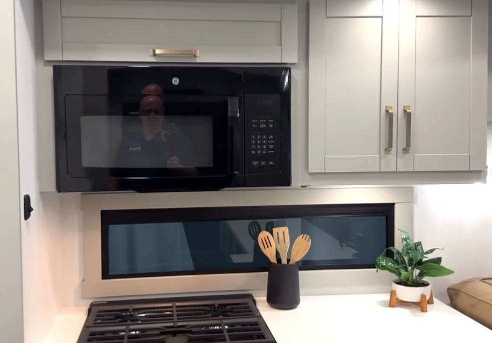 Brinkley Model Z 3200 RV kitchen with GE black microwave, three-burner cooktop, modern gray cabinetry, and horizontal window with decorative utensils and plant.
