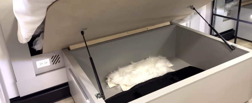 Brinkley Model Z 3200 bedroom under-bed storage compartment lifted open, showing gas-strut supports, spacious interior, and nearby power outlets for added RV convenience.