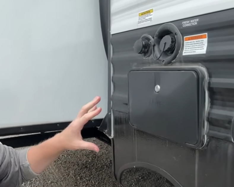 RV exterior utility panel showing fresh water connection, service access door, and plumbing hookups, with a hand pointing to the water system components