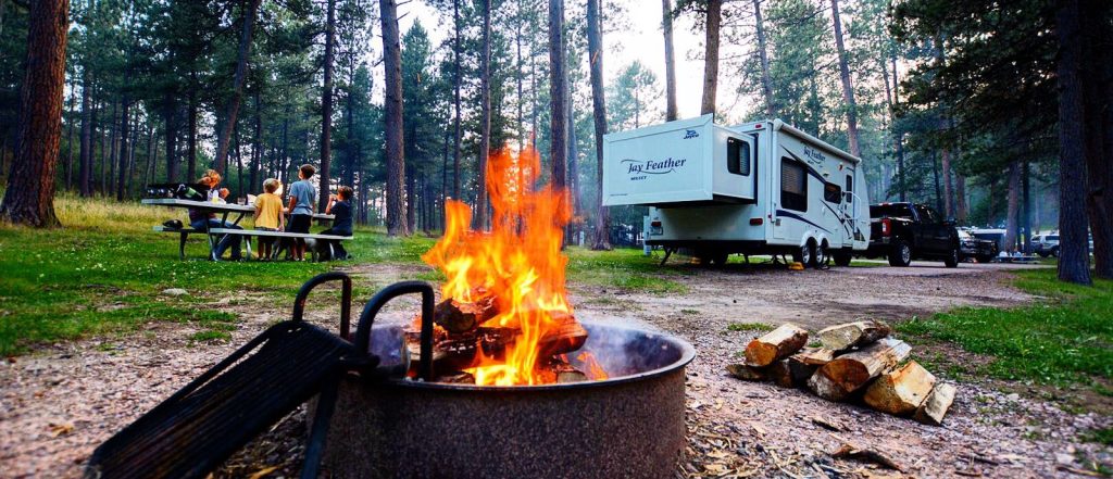 Family enjoying a picnic at a wooded campground near a Jay Feather travel trailer, with a campfire burning in the foreground and a truck parked beside the RV
