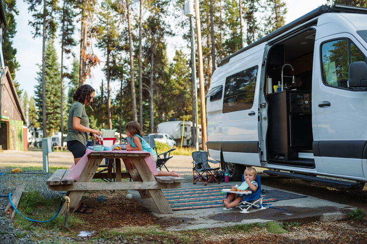 Family camping beside a Class B camper van, preparing food at a picnic table while children play on a mat at a wooded RV campground.