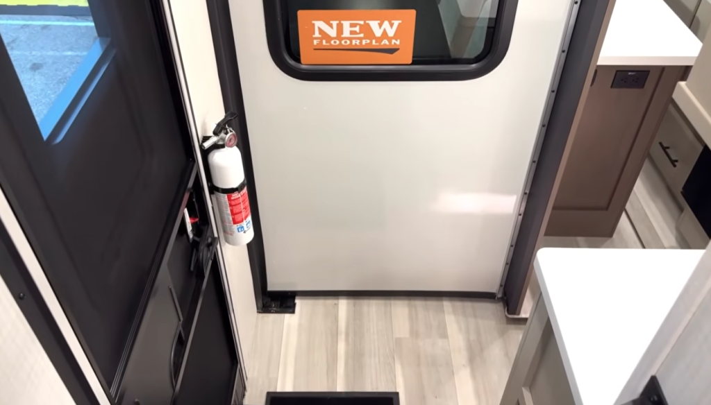 RV interior entryway featuring new floorplan design with solid-surface kitchen countertop, modern cabinetry, fire extinguisher mounted by the door, and durable vinyl plank flooring for easy cleanup and travel durability.