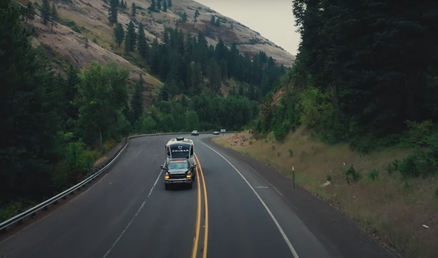 Truck towing a Cougar fifth-wheel RV along a winding mountain highway surrounded by pine trees and rocky hillsides.