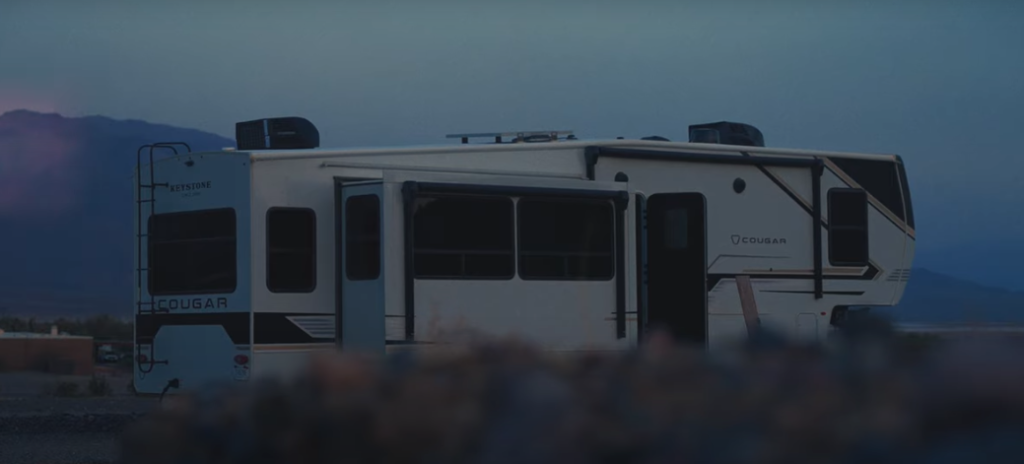 Keystone Cougar fifth wheel RV parked at dusk with slide-outs extended, showcasing the exterior profile and half-ton towable design in a mountain landscape.