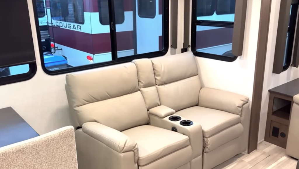 RV theater seating loveseat with plush reclining cushions and built-in cupholders, positioned by large windows in a Keystone Cougar travel trailer living area, showcasing comfortable seating and modern interior design.