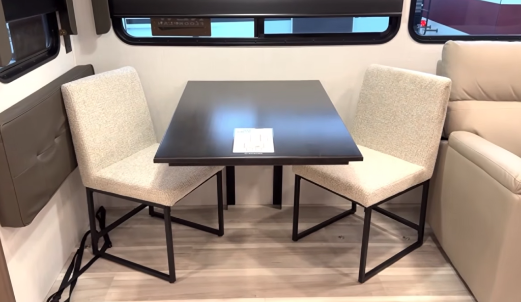 Modern RV dinette with a fixed black dining table and two upholstered chairs, positioned beside a sofa and large window, showcasing a compact eating and workspace area inside a contemporary travel trailer or fifth wheel.