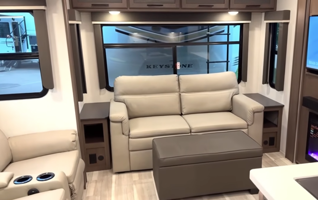 Spacious RV living room featuring a plush theater-style sofa with ottoman, side tables, and large panoramic window, highlighting a comfortable lounge area inside a modern Keystone travel trailer or fifth wheel.