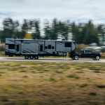 Heavy-duty pickup truck towing a large fifth wheel RV on a highway, showcasing stable towing performance, triple-axle design, and long-distance RV travel through a forested landscape.