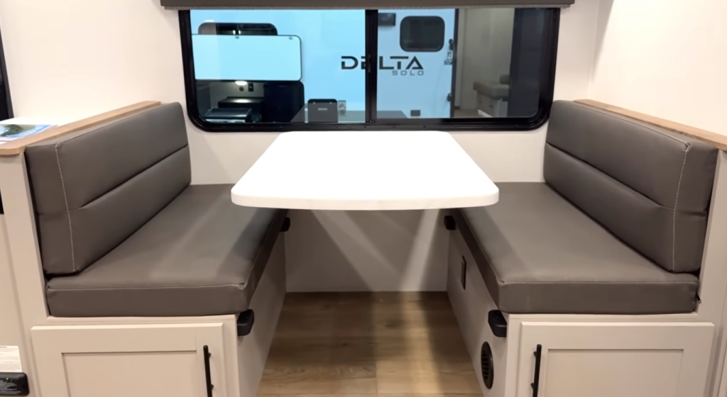 RV dinette with opposing bench seating, cushioned gray vinyl seats, and a rectangular dining table, featuring built-in storage beneath benches and a large window in a lightweight travel trailer interior.