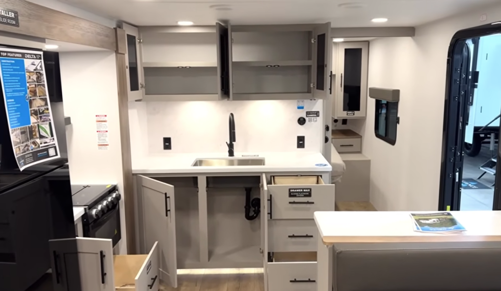Modern RV kitchen with open upper cabinets, deep stainless steel sink, matte black faucet, solid-surface countertops, residential-style drawers, and compact island, showcasing functional storage and clean contemporary RV interior design.