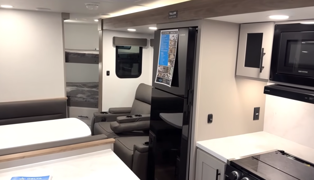 RV interior view showing modern kitchen and living area with black residential refrigerator, microwave, solid-surface countertops, and theater-style recliner seating, highlighting efficient layout and contemporary RV design.
