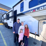 Family of four standing in front of a large fifth wheel RV on a dealership lot, smiling beside their camper.