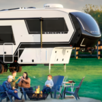 Family relaxing around a campfire beside a Brinkley fifth wheel RV, with outdoor chairs set up on a grassy campsite.