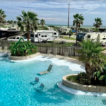 Resort-style swimming pool with in-water lounge chairs, tropical landscaping, and palm trees overlooking a coastal RV resort. Luxury RVs and full-hookup campsites surround the pool area, creating a relaxing vacation atmosphere that combines beachfront camping with upscale resort amenities