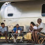 A family relaxing outside their Airstream travel trailer, sitting in camping chairs and talking together at a scenic campsite.