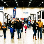 A bright indoor RV show scene in Michigan with families walking between travel trailers and fifth wheels. Warm winter clothing, clean overhead lighting, realistic detail, 16:9 composition.