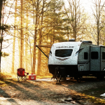 Wide scenic shot of a Michigan state campground in early spring. Mix of pine trees, gravel campsite pad, modern travel trailer with slide pulled out. Soft sunrise light. Include subtle Price Right RV red accent in campsite gear. Clean, documentary style.